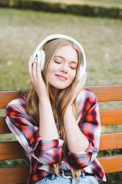 Portrait shot of beautiful girl in casual clothes listening to musing on headphones with closed eyes while sitting on wooden bench in park — Stock Photo