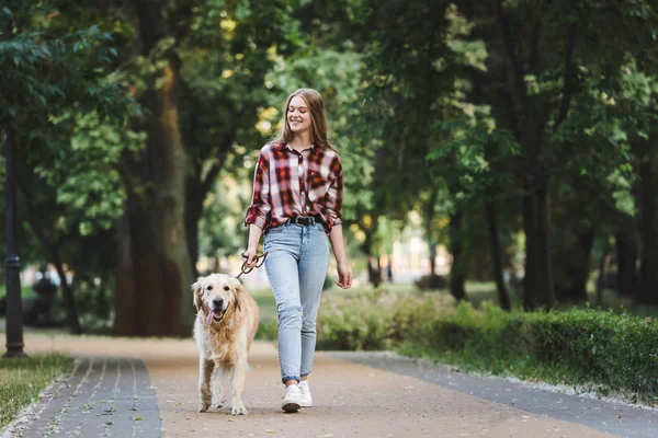 Full length view of beautiful girl in casual clothes waking in park with golden retriever — Stock Photo