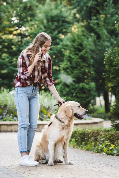 Visão de comprimento total da bela menina em roupas casuais acordando no parque com golden retriever e falando no smartphone — Fotografia de Stock