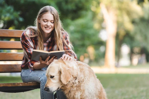 Menina bonita em roupas casuais leitura livro e petting golden retriever enquanto sentado no banco de madeira no parque — Fotografia de Stock