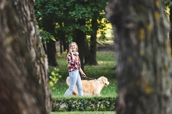 Selective focus of beautiful young girl in casual clothes waking in park with golden retriever and talking on smartphone — Stock Photo