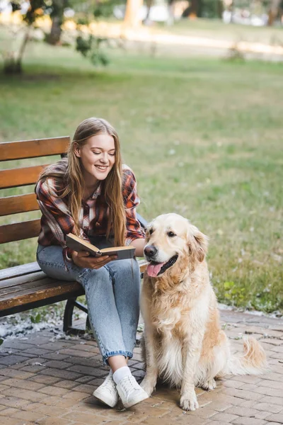 Visão comprimento total da bela menina em roupas casuais leitura livro e petting golden retriever enquanto sentado no banco de madeira no parque — Fotografia de Stock