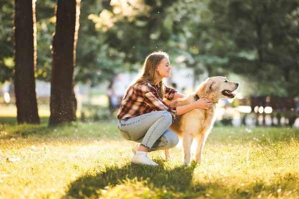 Side view of beautiful girl in casual clothes petting golden retriever while sitting on meadow in sunlight — Stock Photo