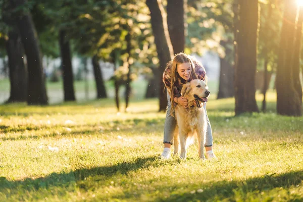 Full length view of beautiful girl in casual clothes hugging golden retriever while standing on meadow in sunlight — Stock Photo