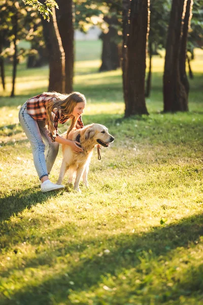 Visão de comprimento total da bela menina em roupas casuais abraçando golden retriever enquanto estava no prado à luz do sol — Fotografia de Stock