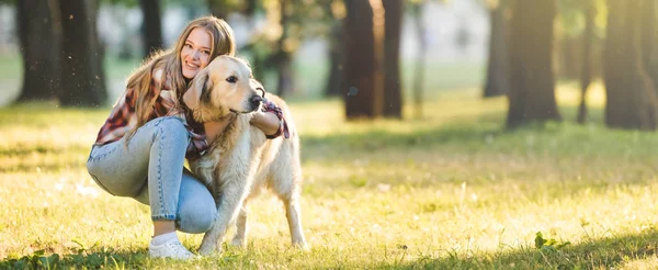 Plano panorámico de hermosa joven en ropa casual abrazando golden retriever mientras está sentado en el prado a la luz del sol - foto de stock