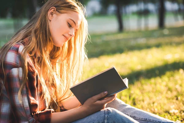 Vista lateral de la hermosa chica en ropa casual sentado en el prado a la luz del sol y libro de lectura - foto de stock