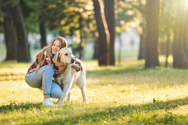 Full length view of beautiful young girl in casual clothes hugging golden retriever while sitting on meadow in sunlight and looking at camera — Stock Photo