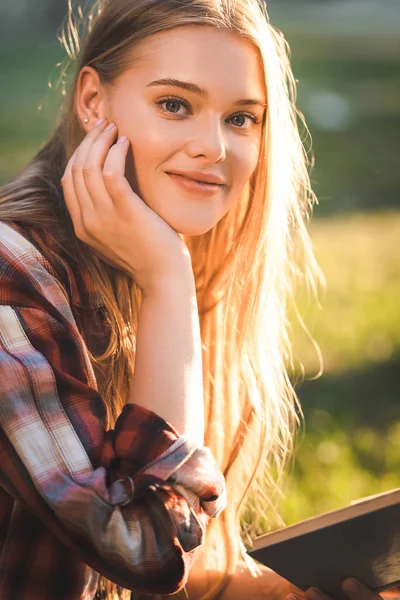 Portrait photo de belle fille en vêtements décontractés assis sur la prairie dans la lumière du soleil, tenant livre et regardant la caméra — Photo de stock