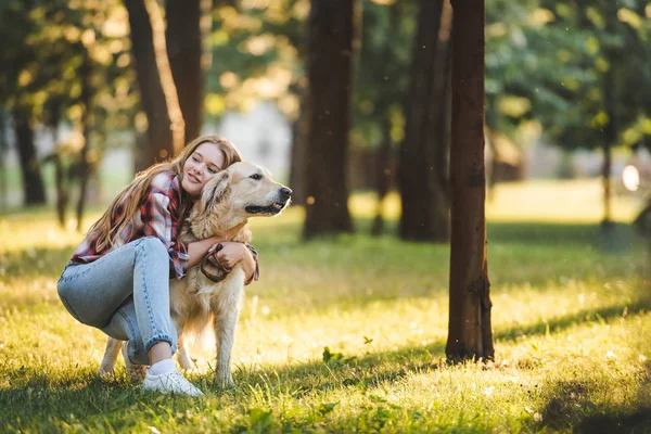 Volle Länge Ansicht der schönen jungen Mädchen in lässiger Kleidung umarmt Golden Retriever, während sie auf der Wiese im Sonnenlicht sitzen und wegschauen — Stockfoto