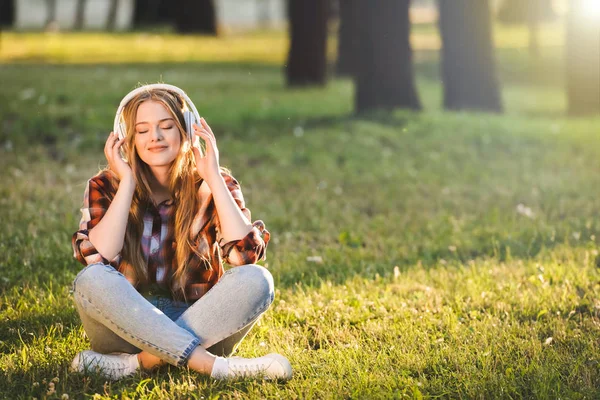 Vista completa de la hermosa chica en ropa casual sentado con las piernas cruzadas en el prado a la luz del sol y escuchar música en los auriculares - foto de stock