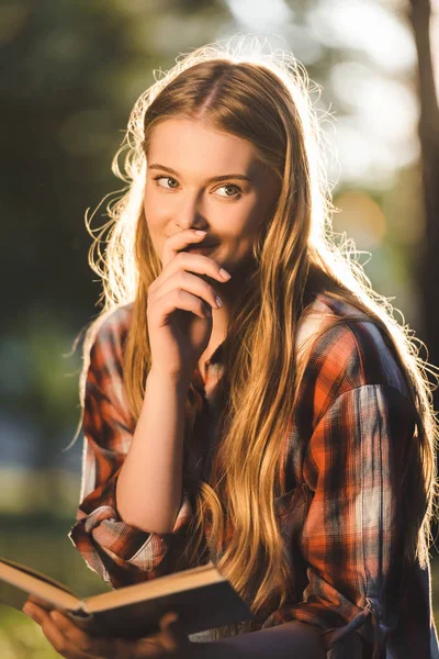 Portrait shot of beautiful girl in casual clothes sitting on meadow in sunlight, holding book and looking away — Stock Photo