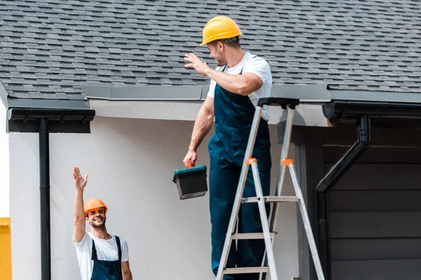 Enfoque selectivo del trabajador saludando mano y mirando a su compañero de trabajo de pie en la escalera con caja de herramientas - foto de stock