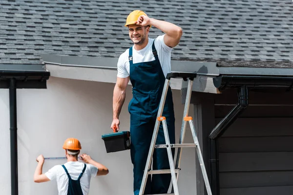 Selektiver Fokus des glücklichen Reparateurs, der Helm berührt und mit Werkzeugkiste auf Leiter neben Mitarbeiter steht — Stockfoto