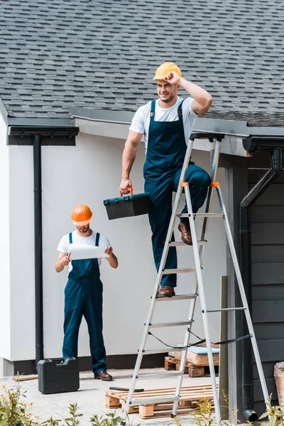 Selective focus of builder touching helmet and standing with toolbox on ladder near coworker — Stock Photo