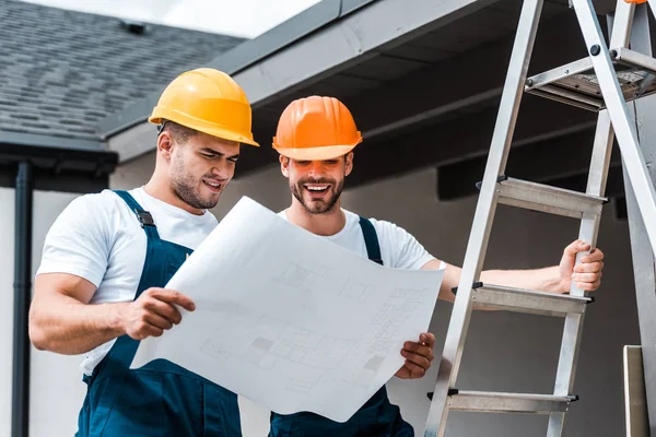 Cheerful builders in helmets looking at paper near ladder — Stock Photo