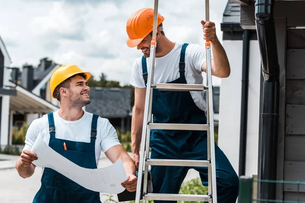 Constructor feliz mirando a su compañero de trabajo en el casco de pie en la escalera - foto de stock