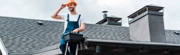 Panoramic shot of happy repairman sitting on roof and holding toolbox — Stock Photo