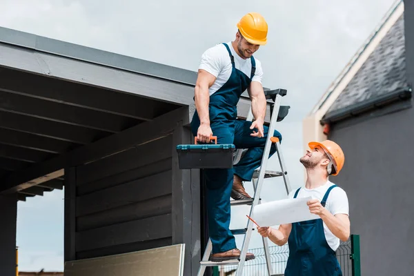 Arquitecto alegre señalando con el dedo en el papel cerca de compañero feliz en el casco - foto de stock