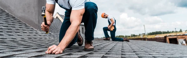 Panoramic shot of handyman holding hammer while repairing roof near coworker — Stock Photo