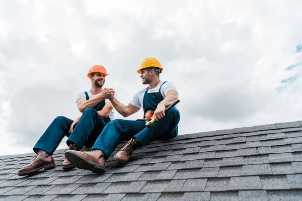 Happy handymen in helmets holding hands while sitting on rooftop — Stock Photo