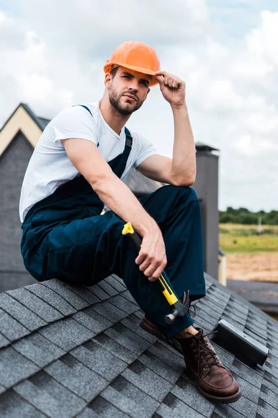 Handwerker mit orangefarbenem Helm sitzt auf Dach und hält Hammer in der Hand — Stockfoto