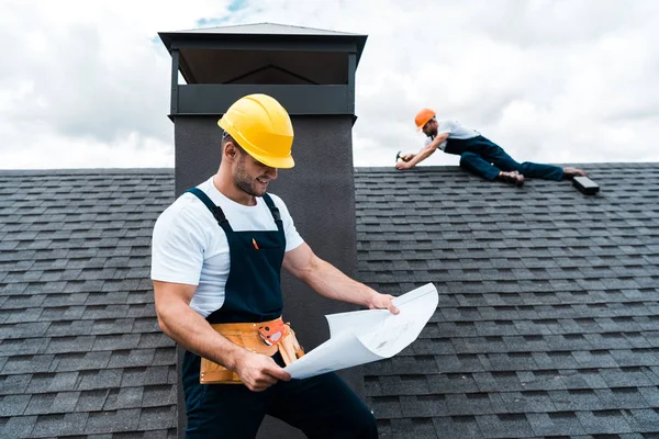 Enfoque selectivo de constructor guapo en casco celebración de anteproyecto cerca de colega reparación de techo - foto de stock