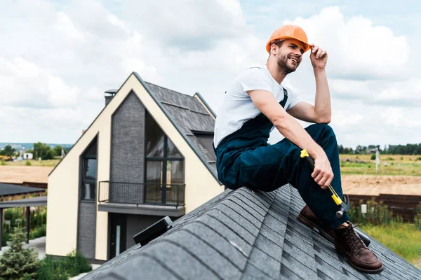 Glücklicher Handwerker mit orangefarbenem Helm sitzt auf dem Dach und hält Hammer in der Hand — Stockfoto