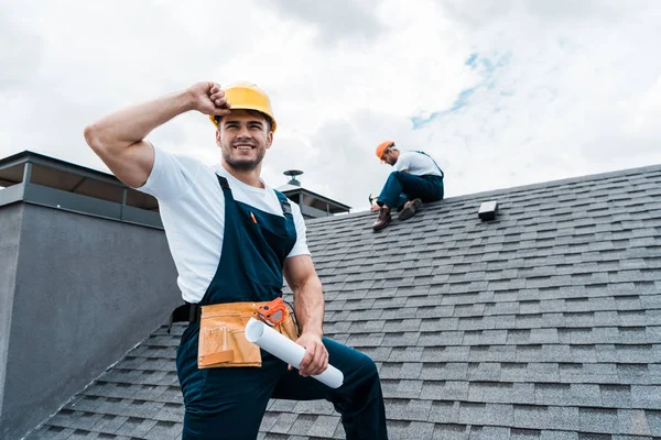 Selective focus of happy repairman holding rolled paper while coworker repairing roof — Stock Photo