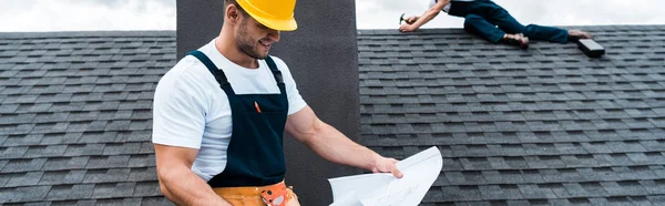 Plano panorámico de constructor guapo sosteniendo plano mientras compañero de trabajo reparación de techo — Stock Photo