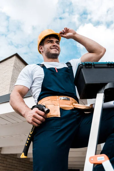Baixo ângulo vista de feliz reparador tocando capacete enquanto segurando martelo — Fotografia de Stock
