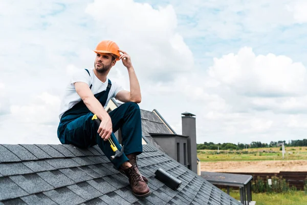 Guapo manitas en casco naranja sentado en el techo y sosteniendo el martillo - foto de stock