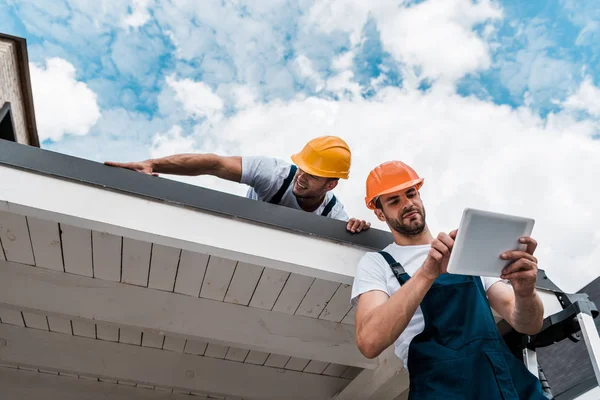 Visão de baixo ângulo de handyman bonito usando tablet digital perto colega de trabalho no telhado — Fotografia de Stock