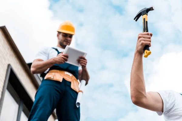 Selective focus of man holding hammer near coworker in helmet using digital tablet — Stock Photo
