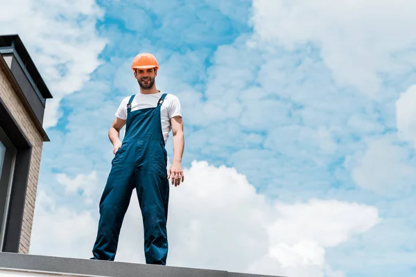 Vista de ángulo bajo de reparador feliz en casco y uniforme de pie contra el cielo azul con nubes - foto de stock