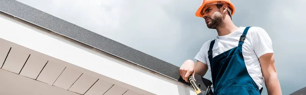 Panoramic shot of repairman in helmet and uniform holding hammer and standing against blue sky with clouds — Stock Photo