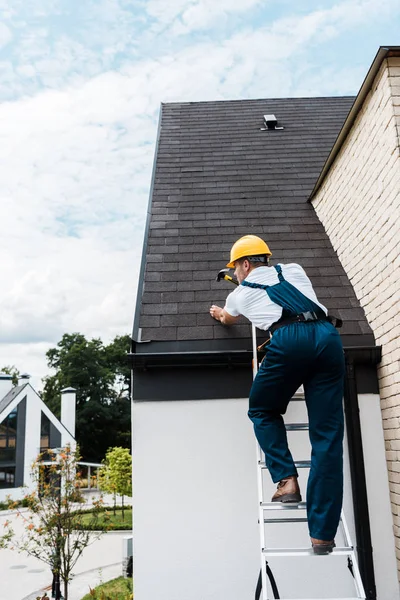 Reparador en uniforme y casco de reparación de techo, mientras que de pie en la escalera - foto de stock