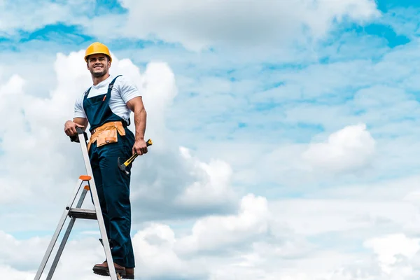 Весёлый ремонтник стоит на лестнице и улыбается на фоне голубого неба с облаками — стоковое фото