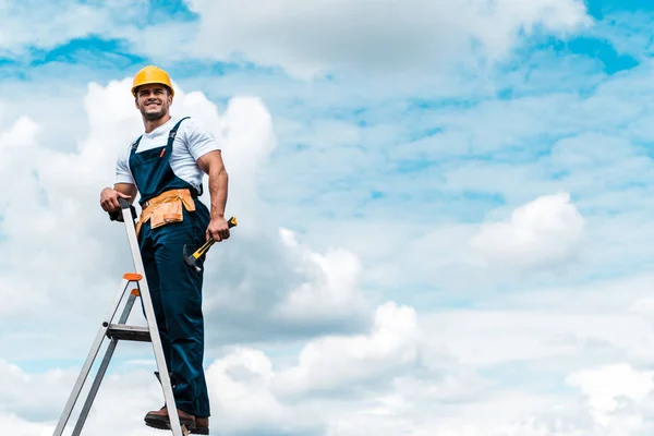 Réparateur joyeux debout sur l'échelle et souriant contre le ciel bleu avec des nuages — Photo de stock