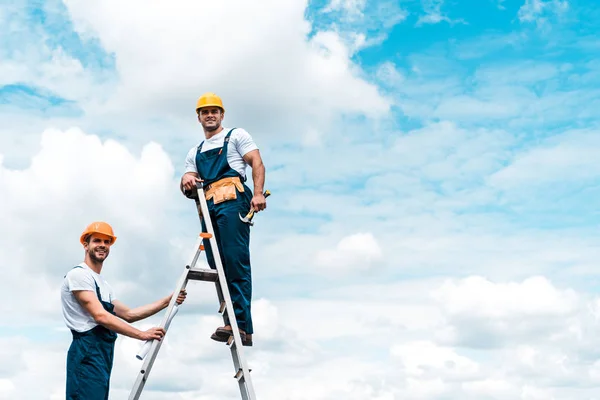 Reparadores felizes em pé na escada e sorrindo contra o céu azul com nuvens — Fotografia de Stock