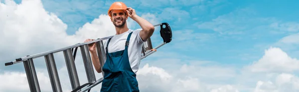 Панорамный снимок веселого ремонтника, держащего лестницу и улыбающегося на фоне голубого неба с облаками — стоковое фото