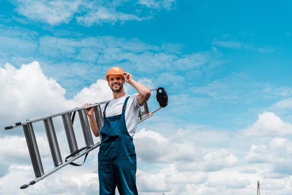 Веселый ремонтник держа лестницу и улыбаясь на фоне голубого неба с облаками — стоковое фото