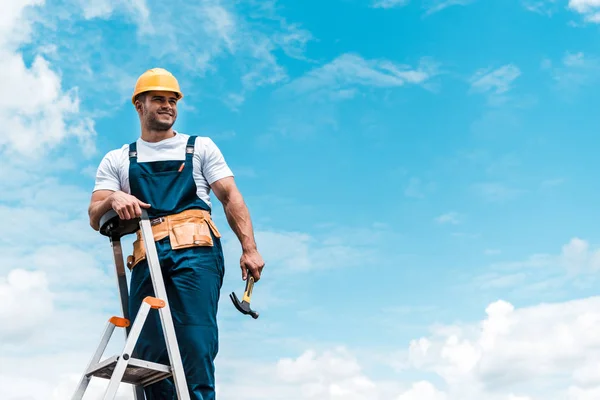 Счастливый ремонтник стоит на лестнице и улыбается на фоне голубого неба с облаками — стоковое фото