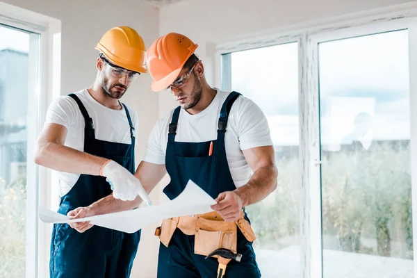 Handsome builders in goggles and helmets looking at blueprint — Stock Photo