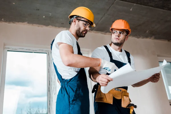 Low angle view of handsome architects in goggles and helmets looking at blueprint — Stock Photo