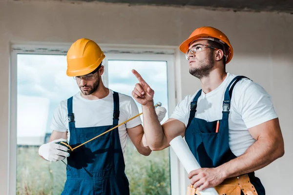 Handsome handyman looking at measuring tape near coworker pointing with finger — Stock Photo