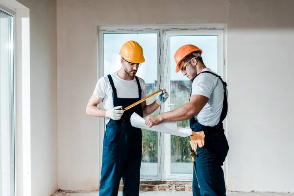 Apuesto constructor mirando plano cerca de compañero de trabajo celebración de cinta métrica - foto de stock