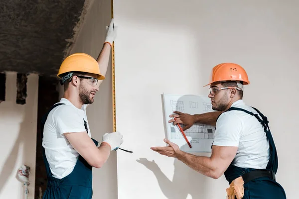 Arquiteto bonito segurando planta perto da parede e olhando para colega de trabalho com fita métrica — Fotografia de Stock