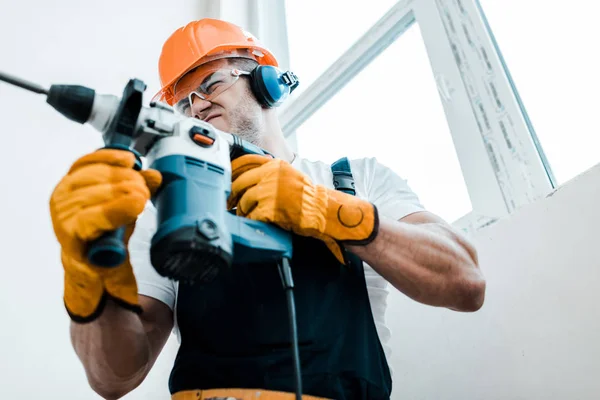 Low angle view of handyman in helmet and yellow gloves using hammer drill — Stock Photo