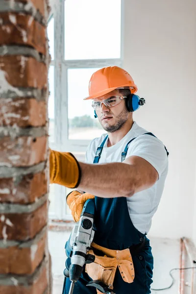 Selektiver Fokus des Arbeiters in Uniform und gelben Handschuhen, der den Bohrhammer in der Hand hält und auf die Ziegelmauer blickt — Stockfoto